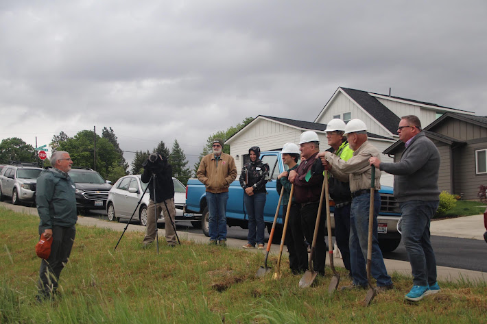 A group of people gathered on a grassy slope for a groundbreaking ceremony under an overcast sky. Several men wearing white hard hats hold ceremonial shovels, while a photographer on a tripod captures the event, and onlookers stand near a blue pickup truck in a residential neighborhood.